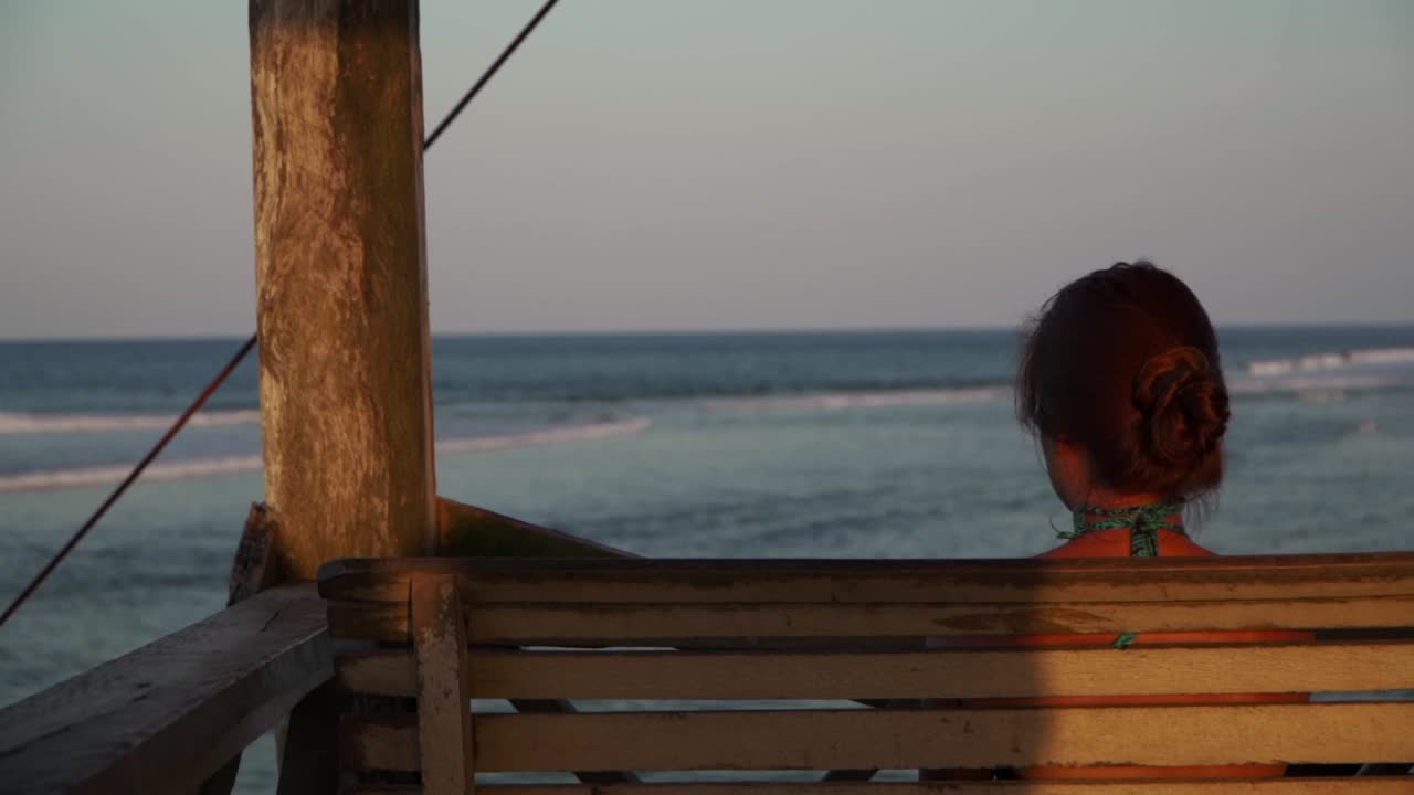 uhd 4k 슬로모로 촬영된 그녀에게 초점을 맞춘 해변의 태평양 충돌의 파도를 보고 있는 젊은 여성의 어깨 너머로 보이는 전망