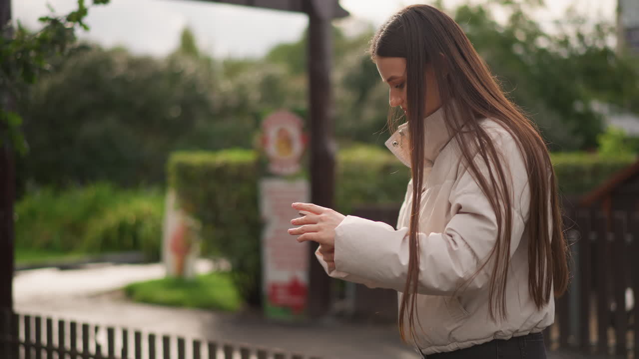 Jovem conferindo o smartphone em plataforma suburbana, usando tranças compridas e jaqueta puffer, rolando mensagens e olhando para cima entre toques, luz nublada, cerca de madeira e vegetação no plano de fundo, casual