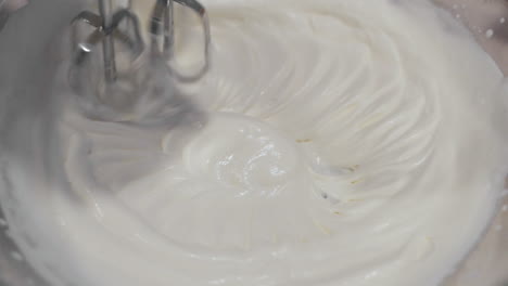 close up macro view of metal mixer stirring and blending sweet white icing in circles in large bowl in slow motion