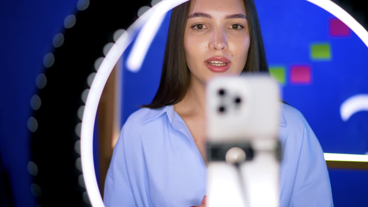 Beautiful Caucasian brunette with long hair stands behind phone on tripod and ring light. Lady talks to camera taking video.