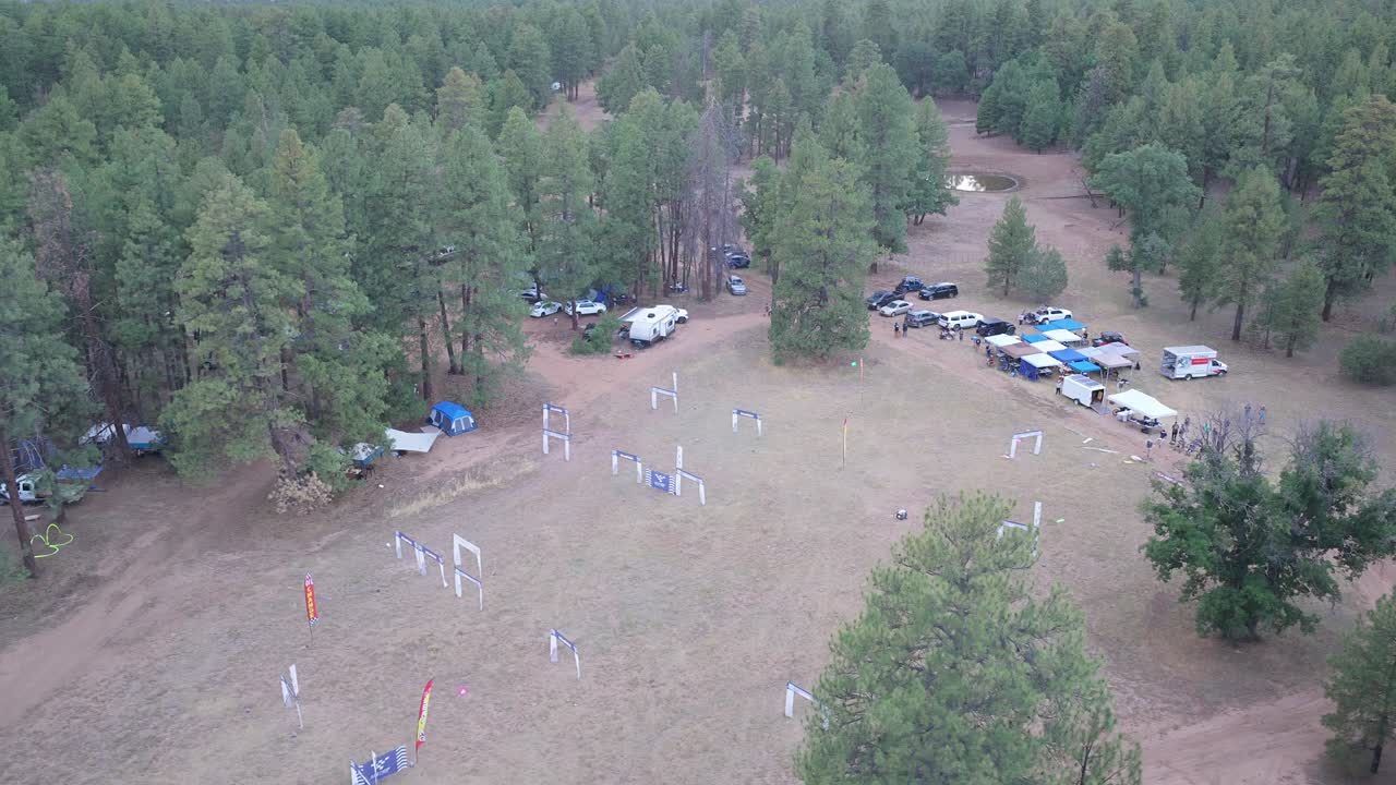 Flygvy över en skogsglänta med en drönarracerbana under gyllene timmen. Tält och campingområde syns bland tallarna. Varmt solnedgångsljus som lyser upp platsen för utomhusevenemanget för motorsport innan skymningen