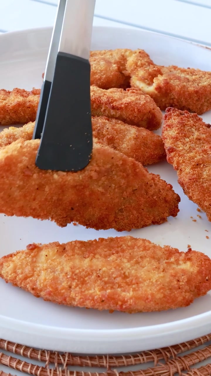 Crispy chicken tenders being arranged with tongs on a white plate, under bright natural lighting