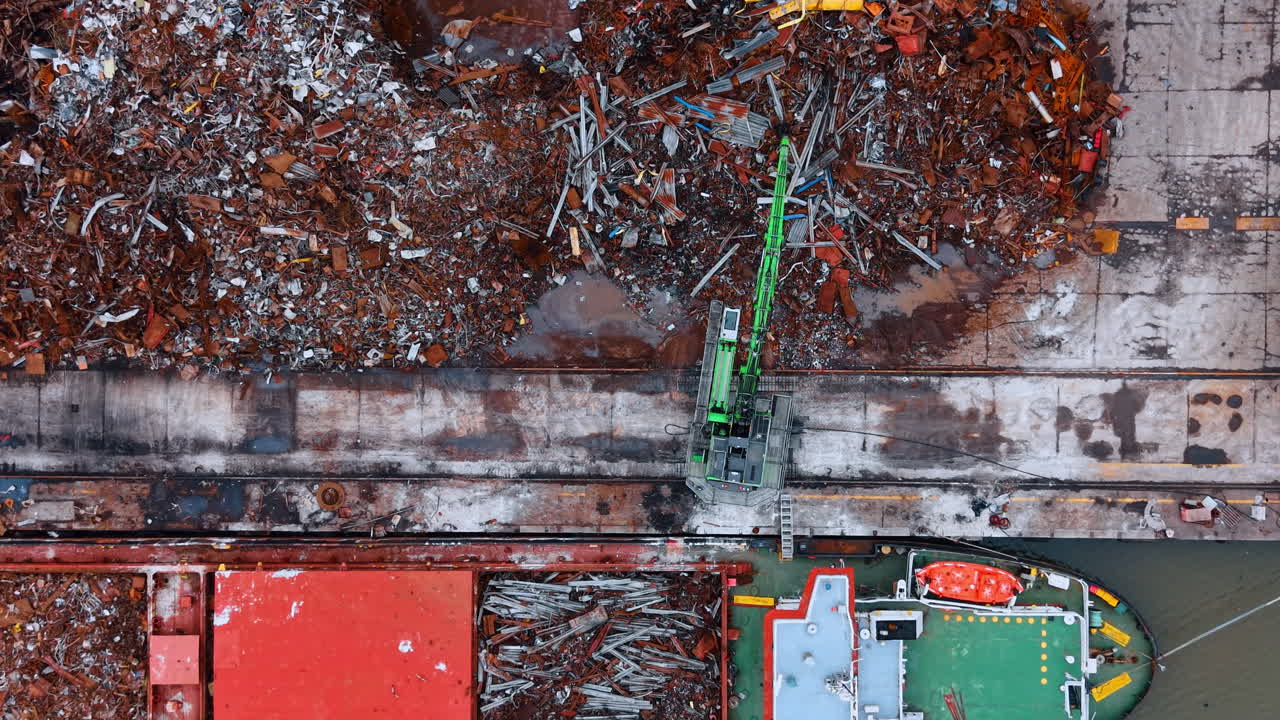 Close up aerial top view of scrap metal loading process. Green hydraulic excavator moving rusty metal scrap from dock pile into bulk carrier cargo hold
