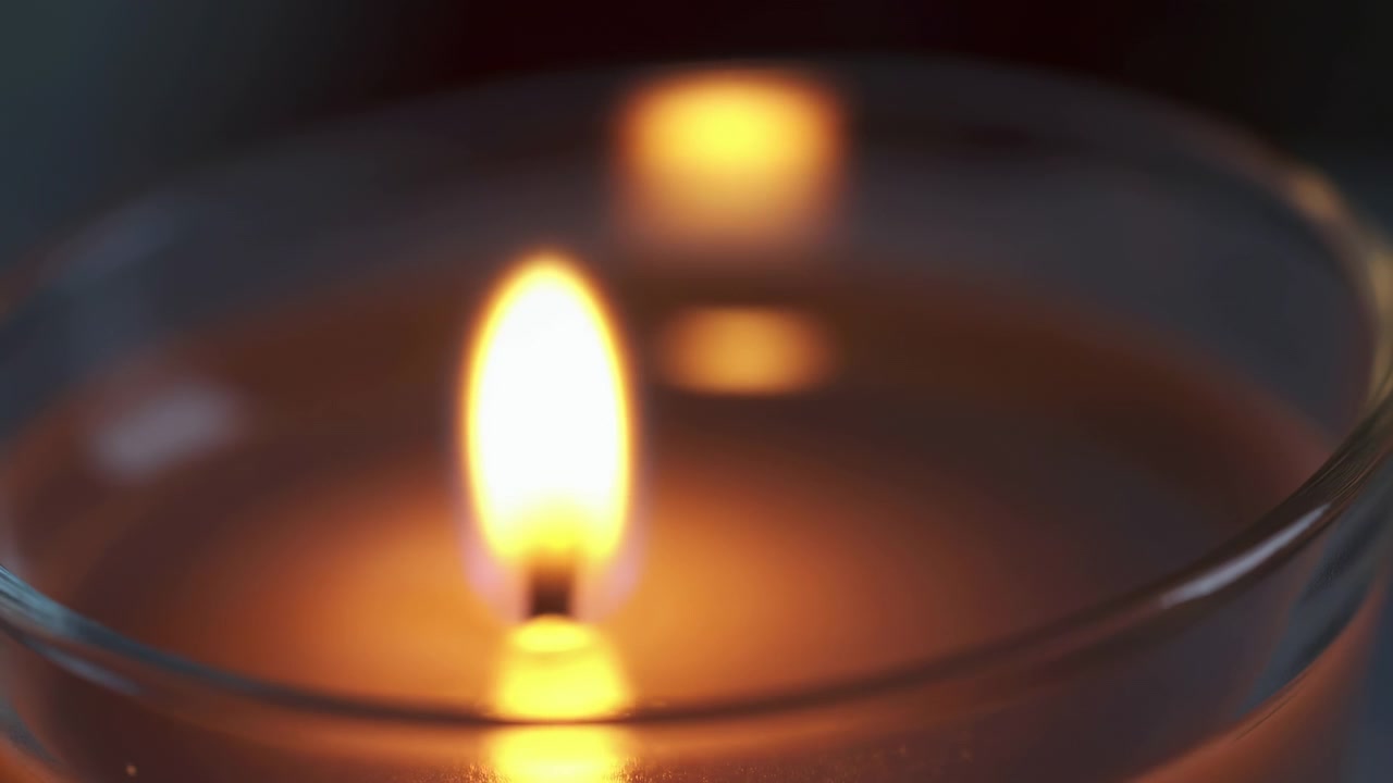 Fiamma di candela in vetro in primo piano, caldo bagliore ambrato con profondità di campo ridotta e morbidi riflessi bokeh sulla cera.