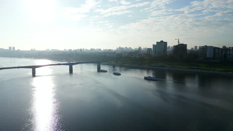 cityscape with a river, bridge, and boats under a bright sky