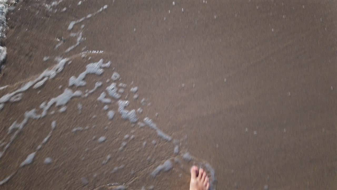 Close up of a man's bare foot touching sea foam while walking on the sandy beach