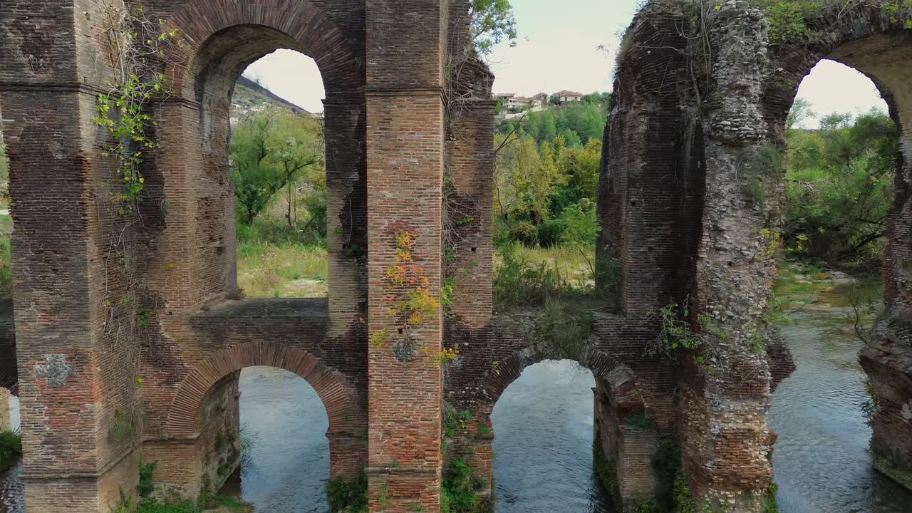 Ripresa drone cinematografica con tracking verso destra lungo l'antico Acquedotto Romano di Nicopolis, caratterizzato dagli archi ripetuti in mattoni rossi sul fiume Louros in Epiro, Grecia.