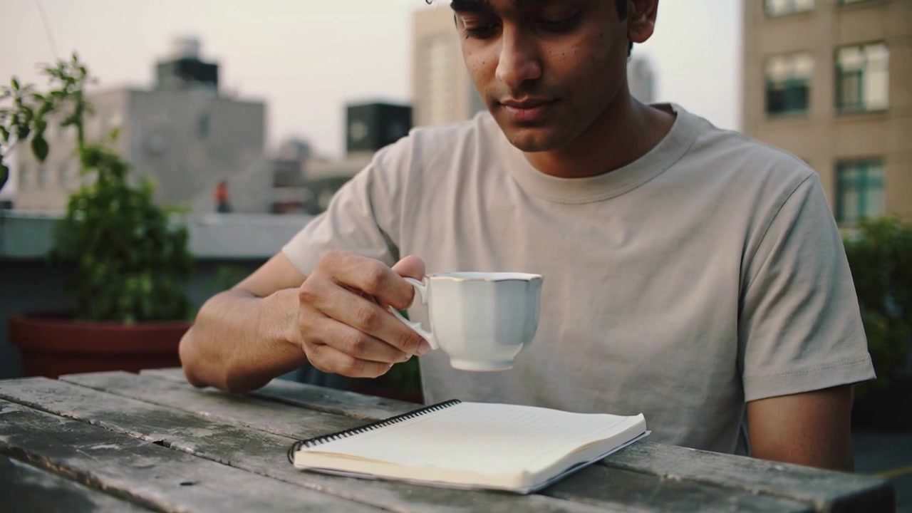 Un homme en t-shirt et casquette sombre levant une tasse de thé blanche, un carnet attirant son regard sur une terrasse sur le toit.