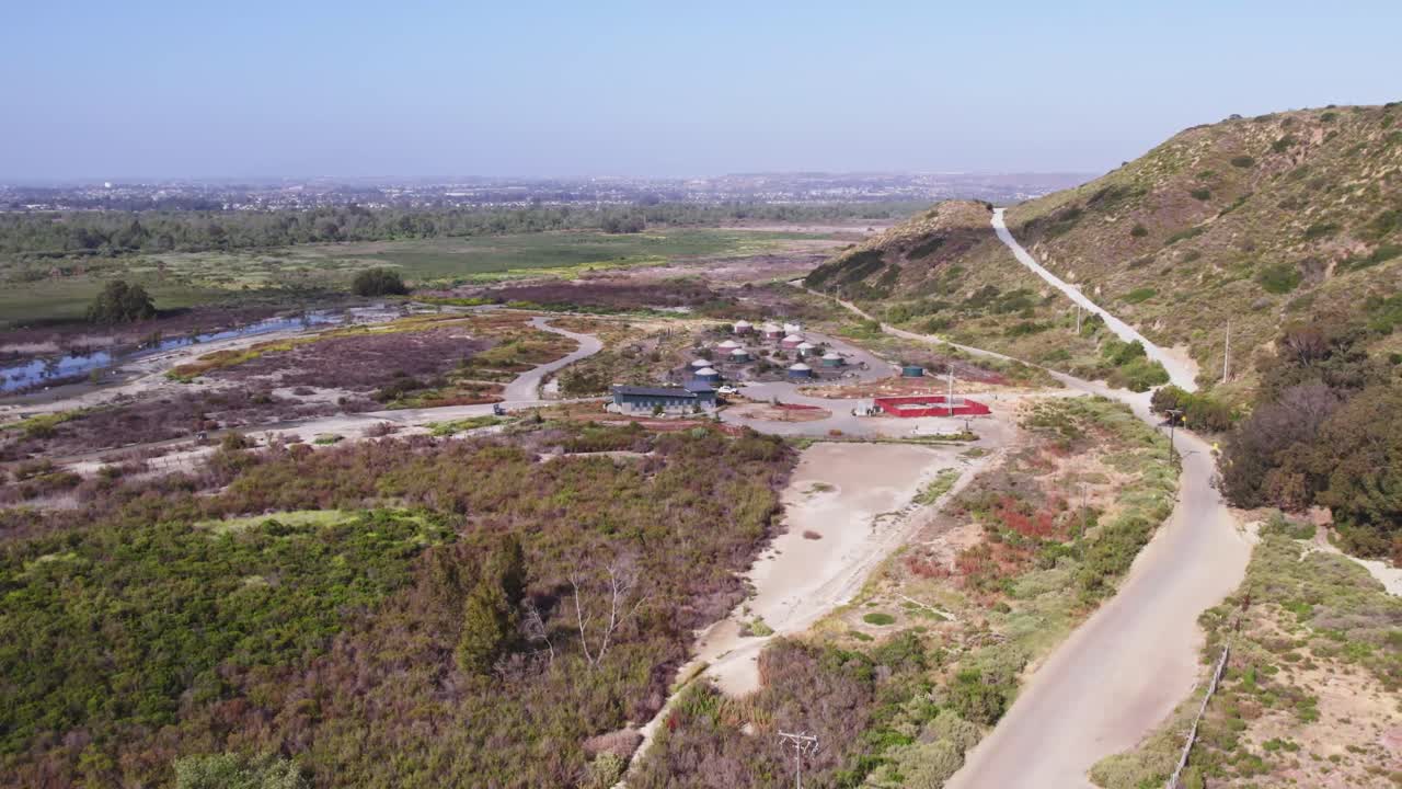 Aerial view over marshland meeting dry border terrain, with a cluster of buildings nestled at the edge of a hillside and a snaking access road.