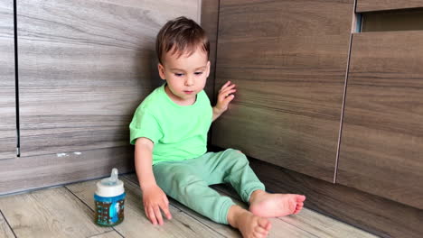 lovely caucasian baby sits comfortably on the floor. toddler touches the floor and then waves his feet. bottle with pacifier is next to the boy.