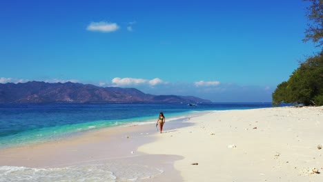 happy tourist woman in green bikini walking on the white sand beach holding snorkeling geatr with cloudy blue sky above