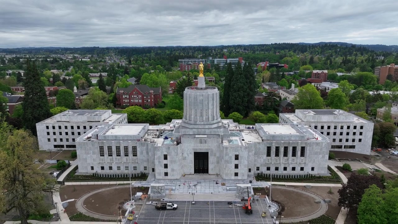 Oregon State Capitol-bygningen i Salem med hvid marmorfacade og Art Deco-kuppel med gylden statue omgivet af frodige grønne træer under overskyet himmel om foråret i USA. Luftfoto, bred stigning.