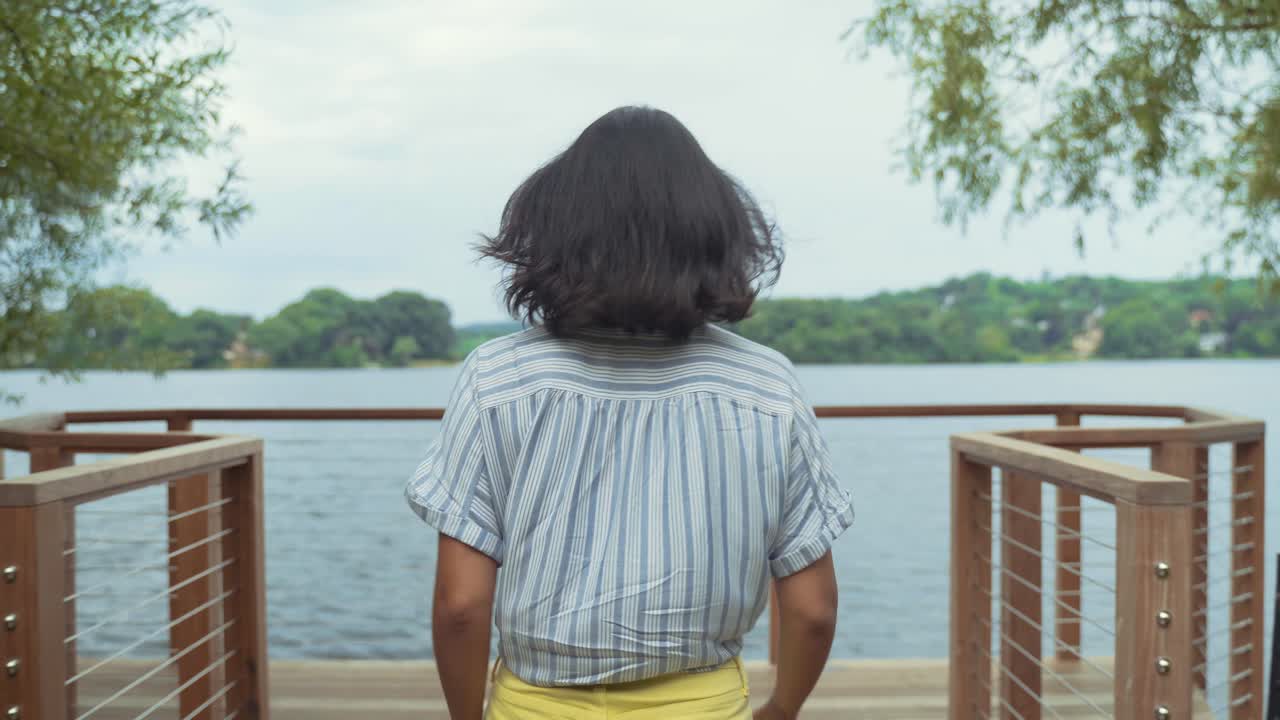 Young Hispanic Woman Walking On Dock By Lake To Get Some Fresh Air, Leans On Bannister To Enjoy Breeze