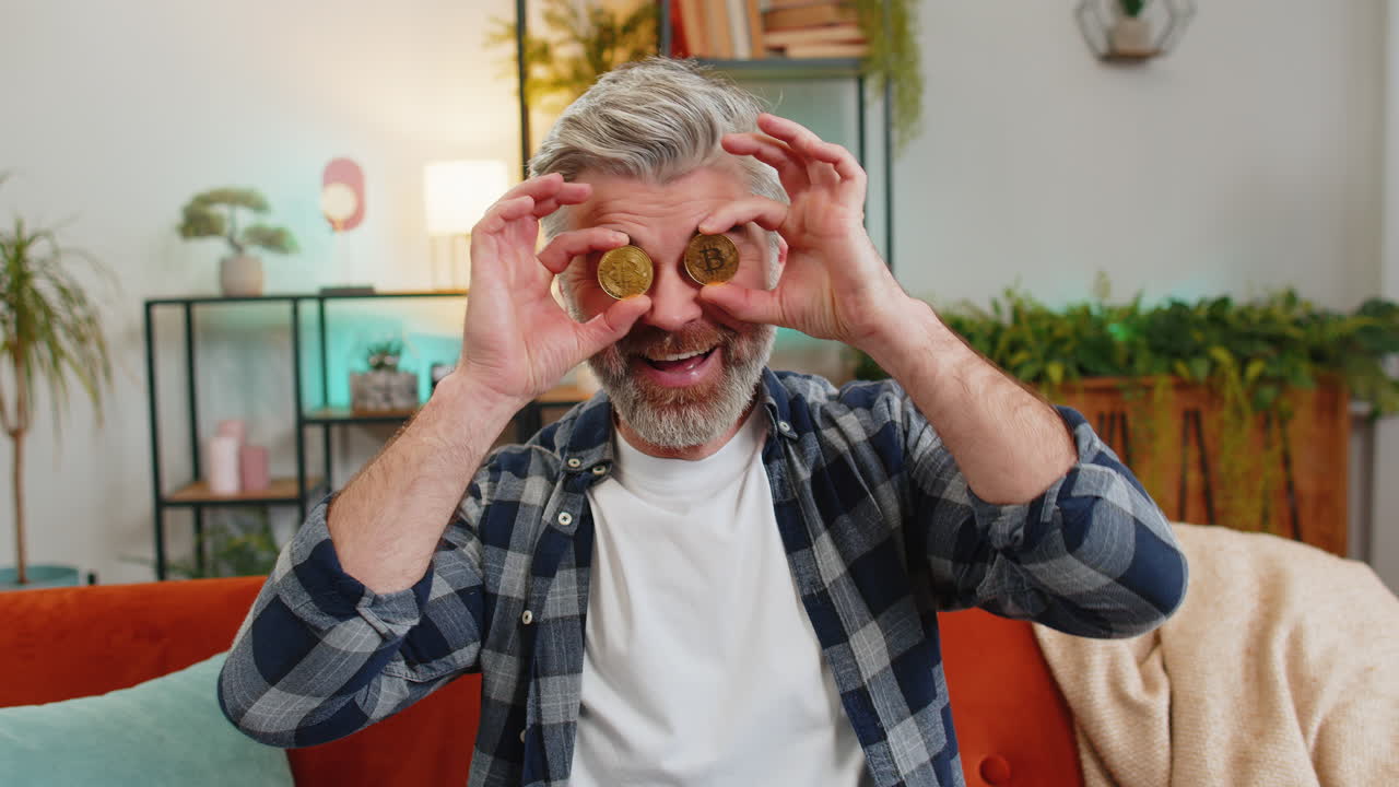 Happy middleaged man successful stock trader holds covering eyes with golden bitcoins at home room