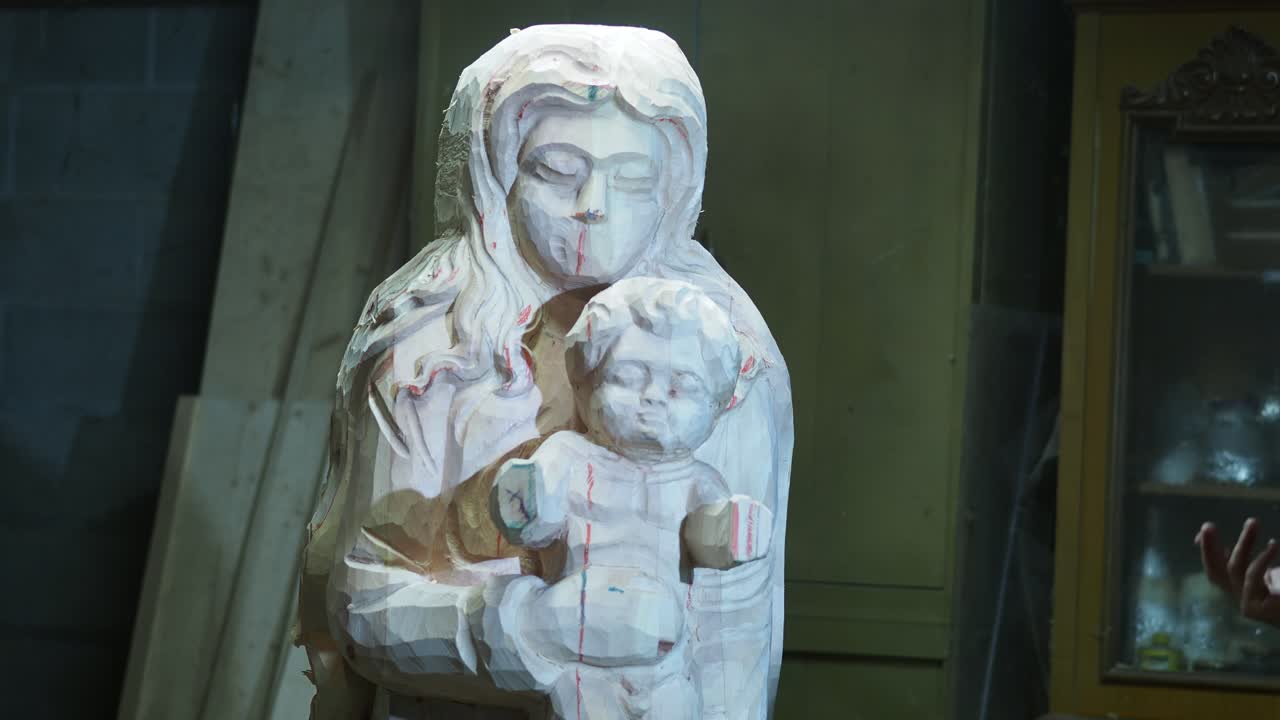 A partially carved wooden statue of the Virgin Mary holding the Child is shown in progress. The artisan uses hand tools to shape the figure, leaving visible chisel marks and guide lines