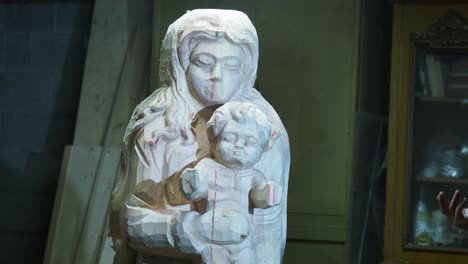 a partially carved wooden statue of the virgin mary holding the child is shown in progress. the artisan uses hand tools to shape the figure, leaving visible chisel marks and guide lines