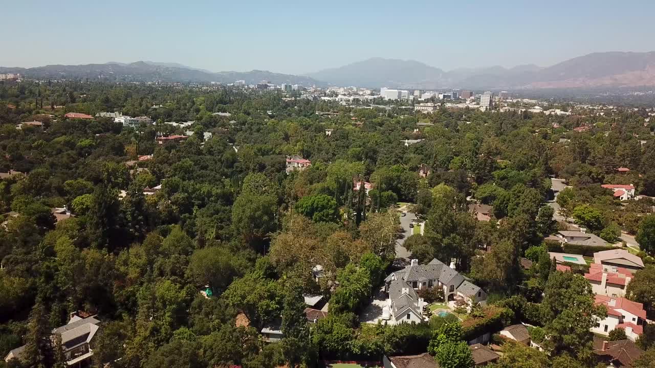 Wide aerial pullback from above an upscale Pasadena neighborhood, retreating to reveal dense tree canopy, large single-family homes (tennis court visible), and the San Gabriel Valley skyline