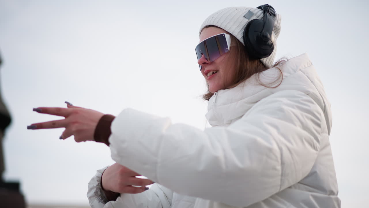 Side view of girl standing beside statue in park rolling hands with joyful energy wearing white coat beanie visor headphones against snowy urban setting capturing playful motion
