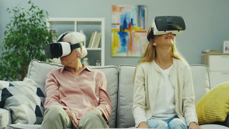 young teenager girl and her senior grandmother in vr glasses having vr headset and looking around as seeing virtual world in the living room. inside.