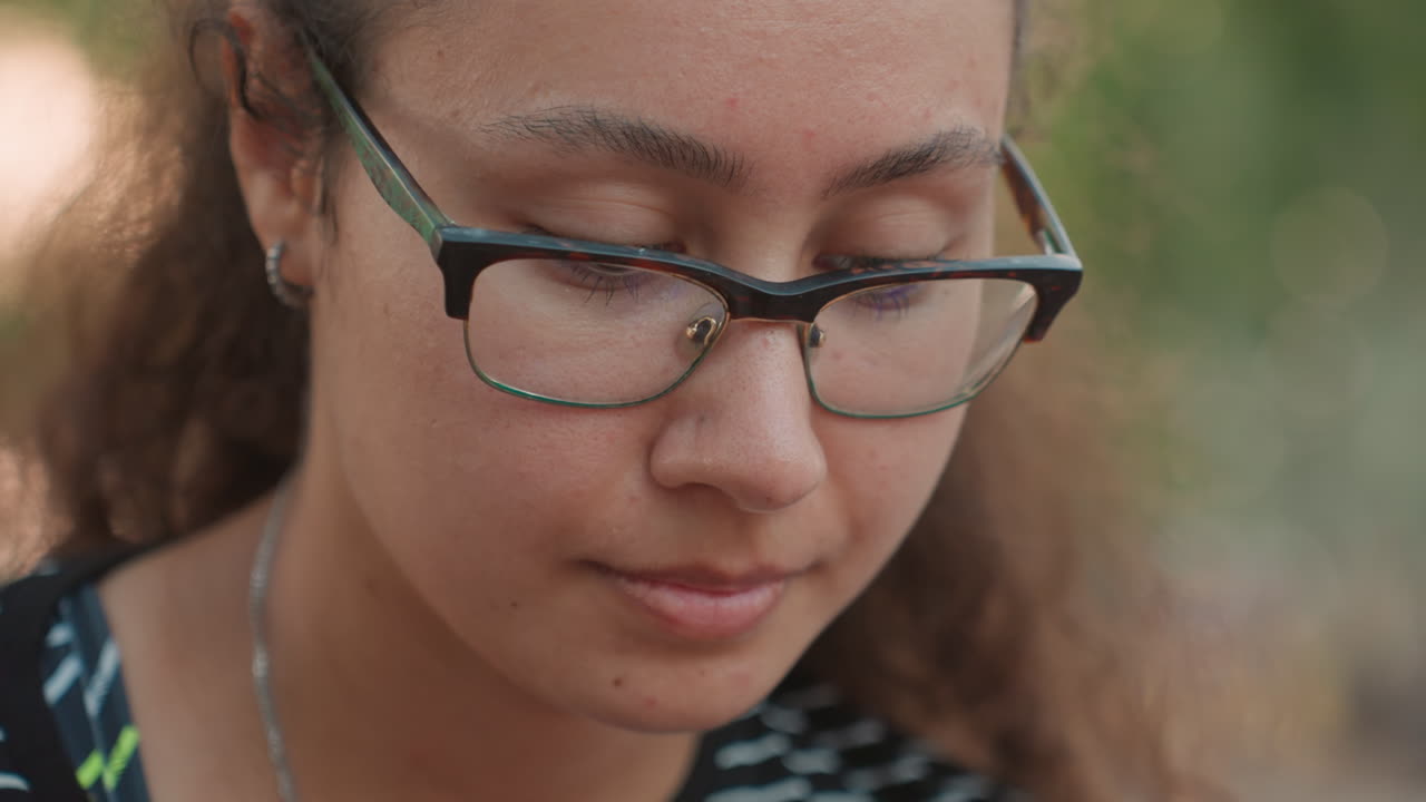 Giovane donna con occhiali che guarda in basso in un ambiente di parco all'aperto, sfondo bokeh morbido, capelli ricci raccolti, top a righe e collana delicata, sottile riflesso sulle lenti, sguardo che si sposta verso l'alto