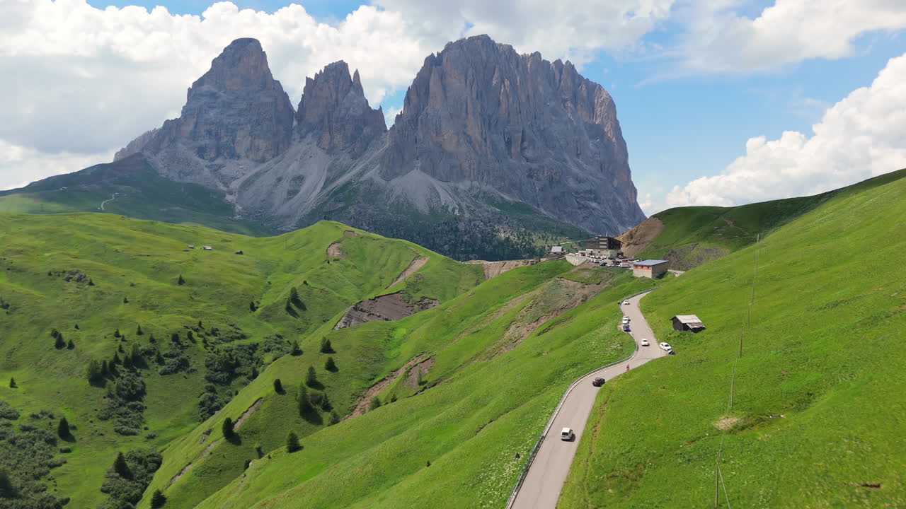 Aerial view of winding alpine road with cars moving towards dramatic Dolomites peak, captured in summer with green slopes and clear sky