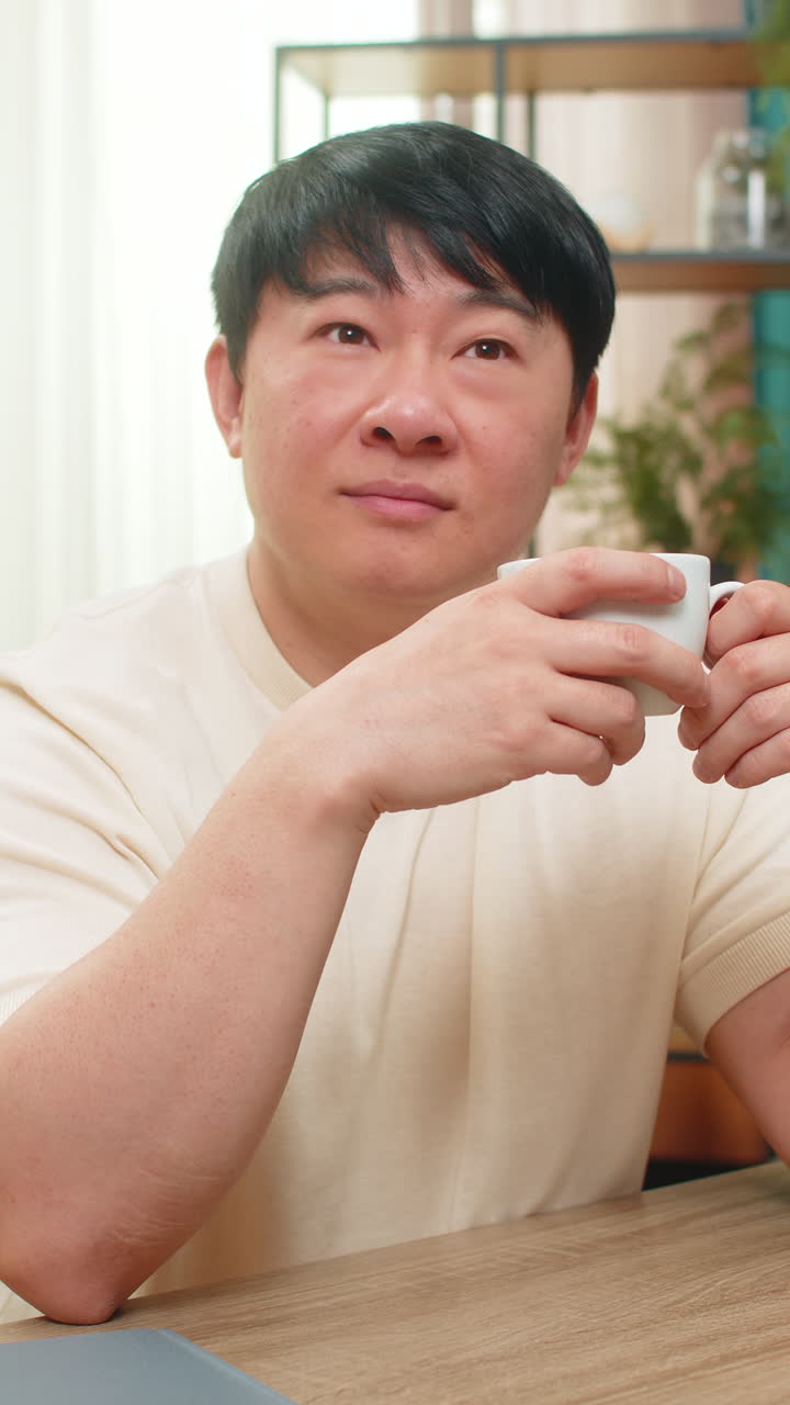 Asian man holding cup of coffee gazing out window thoughtful soft mood enjoying quiet reflection