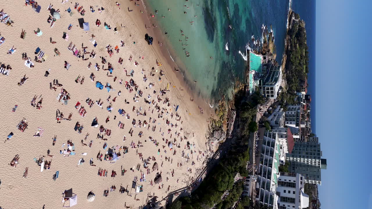 Drone footage over Bondi Beach’s ocean pool, revealing the vibrant blue waters and scenic coastline. Vertical aerial footage starting on the beach flying over pool.