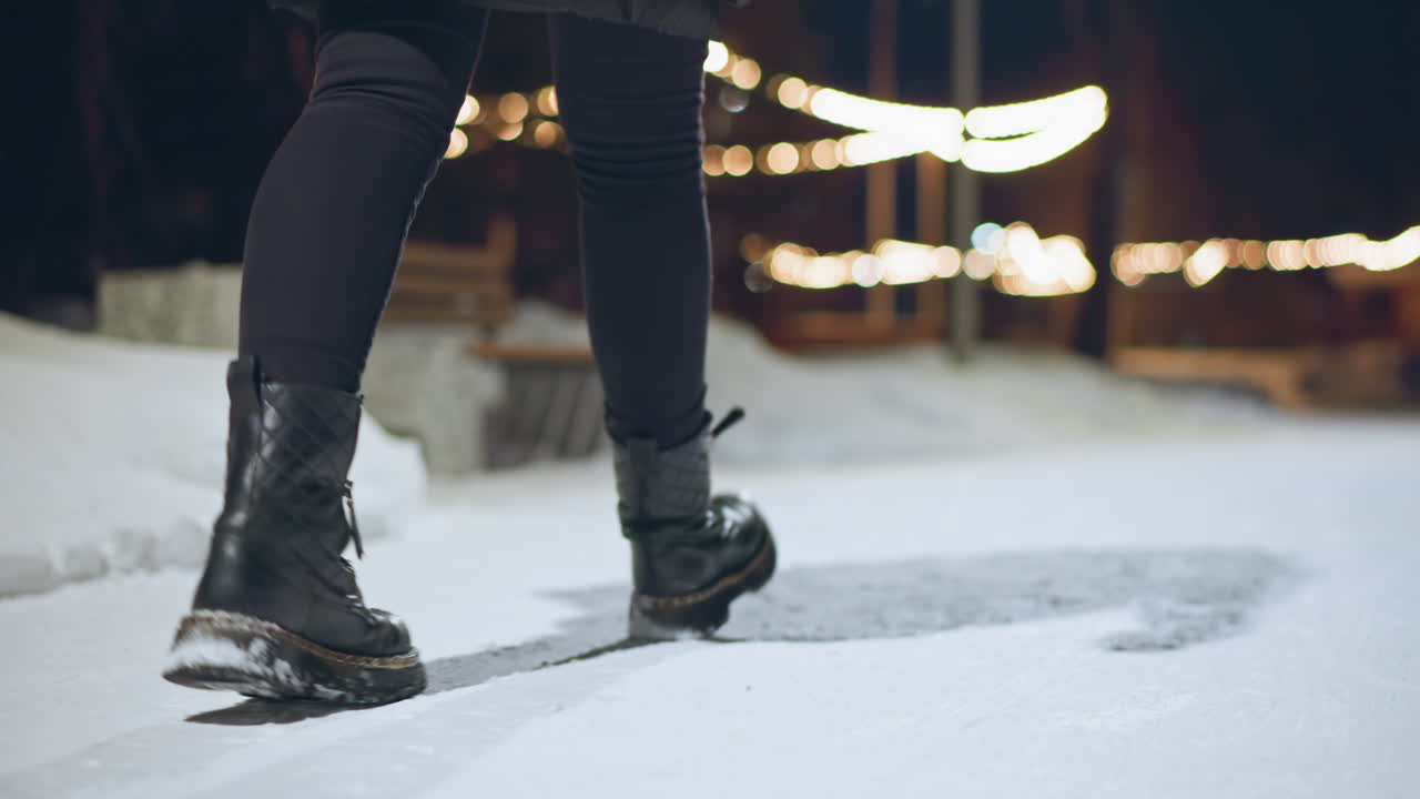 Primer plano de una persona con botas negras de invierno caminando por un sendero nevado de noche con luces festivas brillantes al fondo, lo que crea un fuerte contraste del calzado oscuro contra el suelo helado brillante.