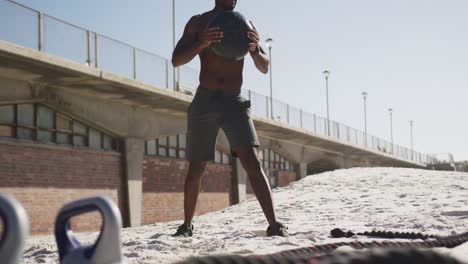 focused african american man lifting ball, exercising outdoors on beach