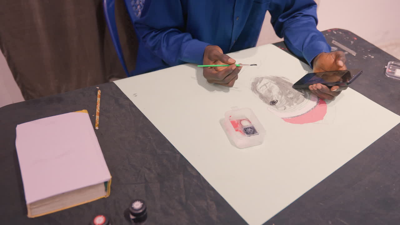 Black artist refining portrait using phone reference, tabletop studio scene with notebook and paint jars, steady brushwork adding midtone shading and pink crescent, professional commission vibe, hands