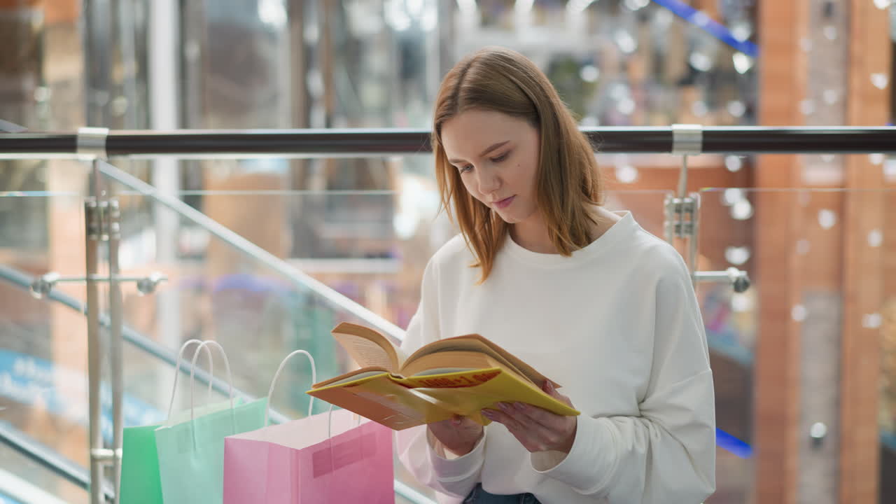 mujer joven sentada en un centro comercial moderno leyendo libro amarillo pensativo con bolsas de compras cerca, barandillas de vidrio contemporáneas y reflejos de luz suave