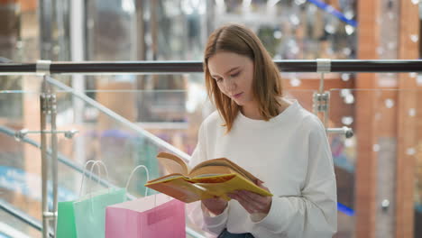 mujer joven sentada en un centro comercial moderno leyendo libro amarillo pensativo con bolsas de compras cerca, barandillas de vidrio contemporáneas y reflejos de luz suave