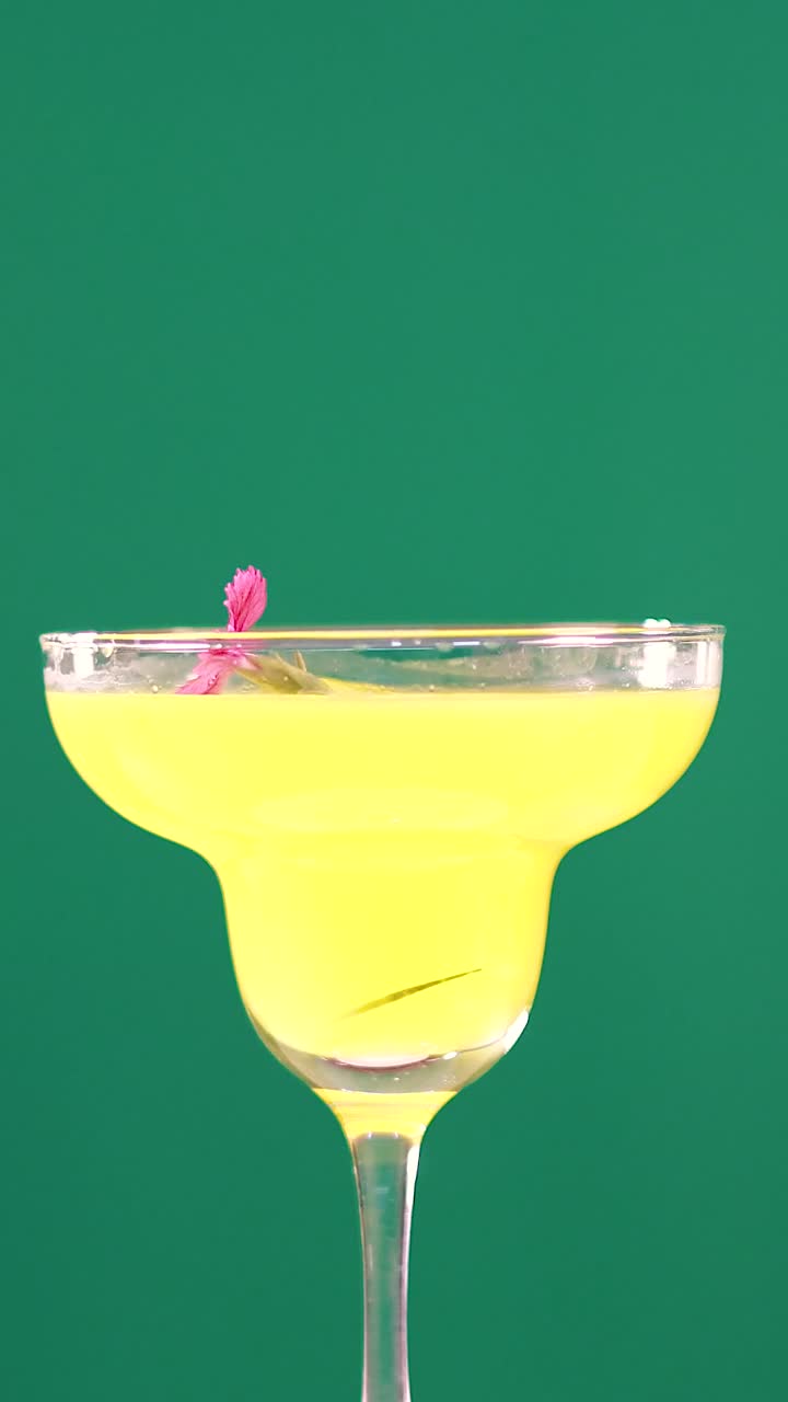 Um coquetel margarita vibrante amarelo em taça com uma guarnição de flor rosa contra um fundo verde.