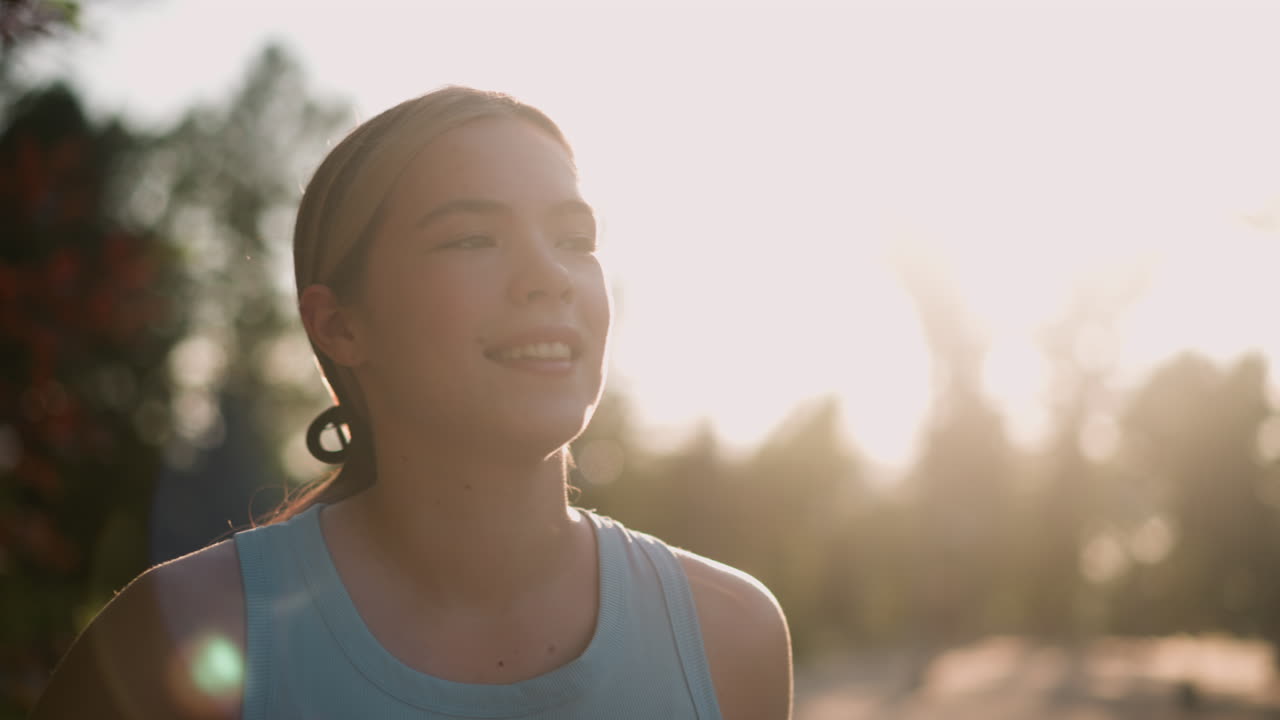 Close-up of young lady smiling with sun glowing around her face, creating a warm, radiant backlit effect, she appears serene and joyful, hair tied back, with blurred trees in background