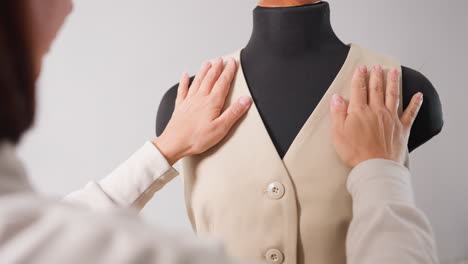back view garment maker adjusts beige sleeveless vest on black mannequin, hands smoothing shoulders, aligning seams, ensuring drape during meticulous fitting inside minimalist fashion studio
