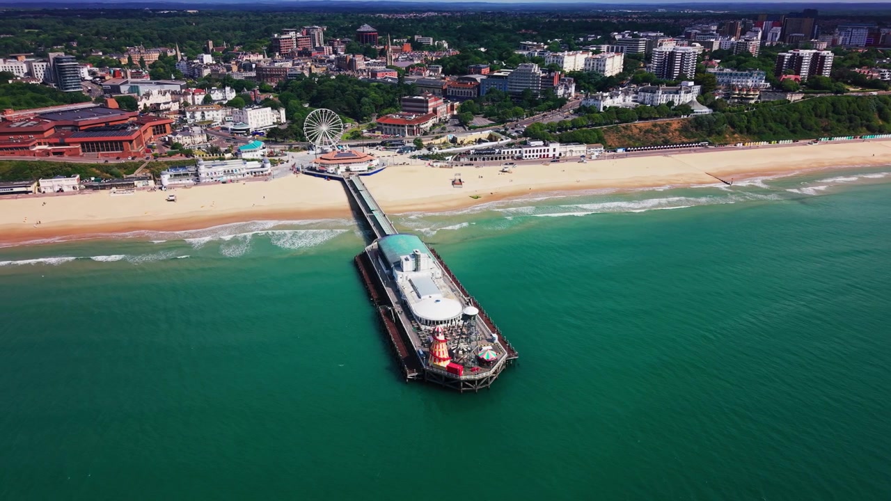 Luftoptagelser med drone af Bournemouth Pier, der strækker sig fra sandstranden ud i turkist hav, med bølger, kystnære attraktioner, pariserhjul, hoteller, klipper og kystbyens skyline.