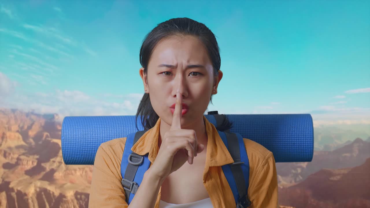 Close Up Of Asian Female Hiker With Mountaineering Backpack Looking At Camera And Making Shh Gesture While Traveling At The Top Of Mountain