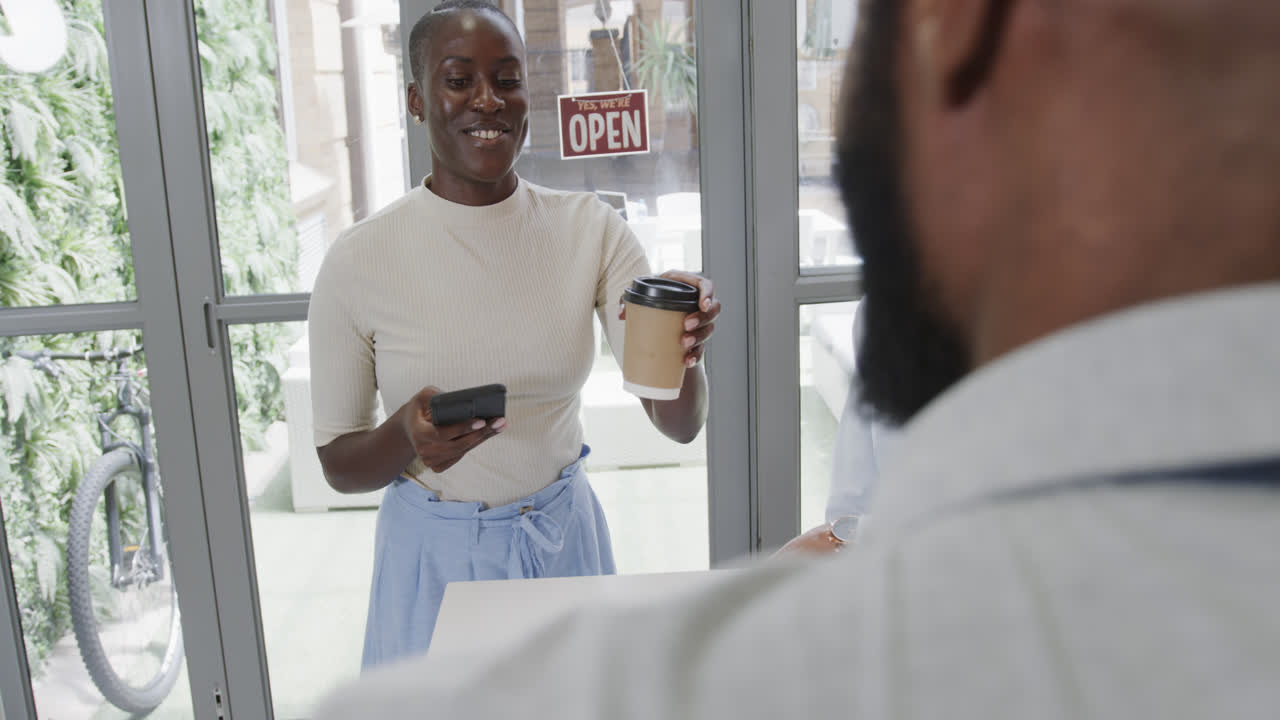 una mujer afroamericana sonriente pagando por un café para llevar en un café con un teléfono inteligente, en cámara lenta