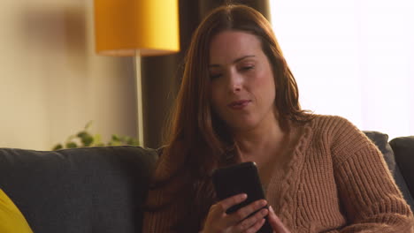 woman sitting on sofa at home using mobile phone to check social media message and read news article 1