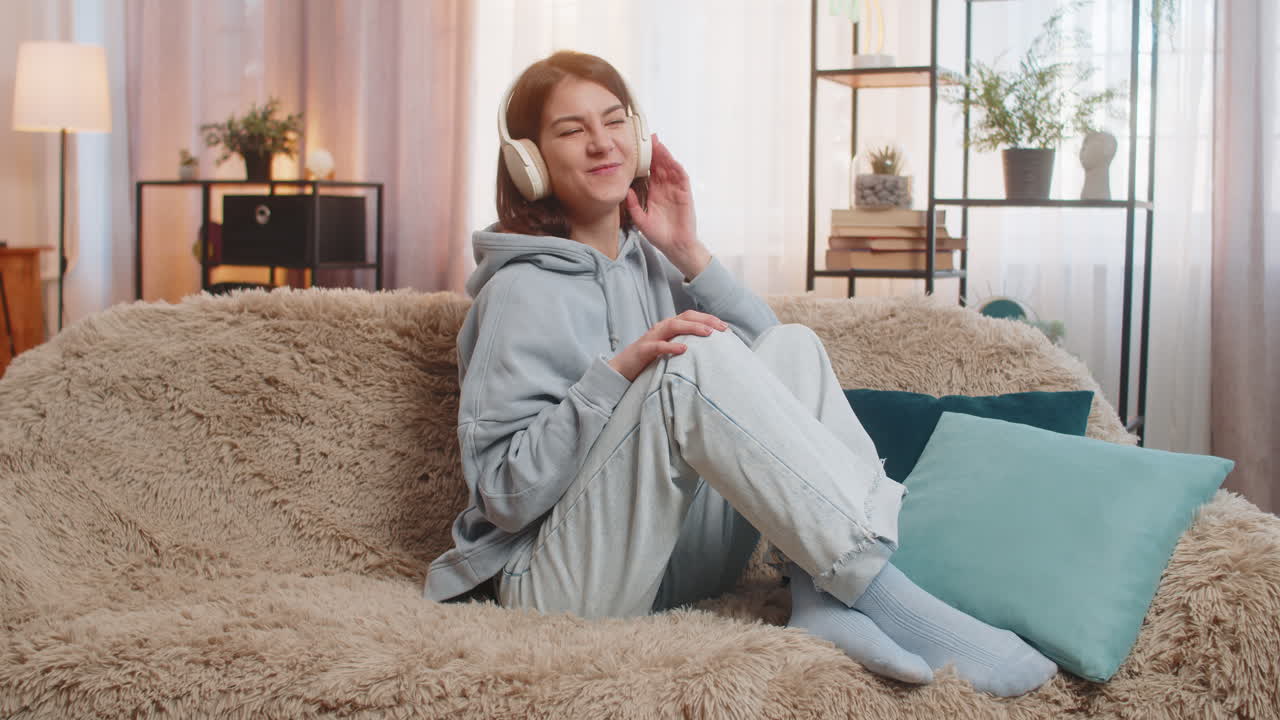 Joyful young woman listening to music in wireless headphones moving to the rhythm sitting on sofa