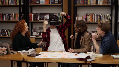 The-boy-in-plaid-shirt-is-wearing-virtual-reality-glasses-in-the-college-library-among-his-classmates.-The-concept-of-modern-technologies-and-virtual-life-in-education