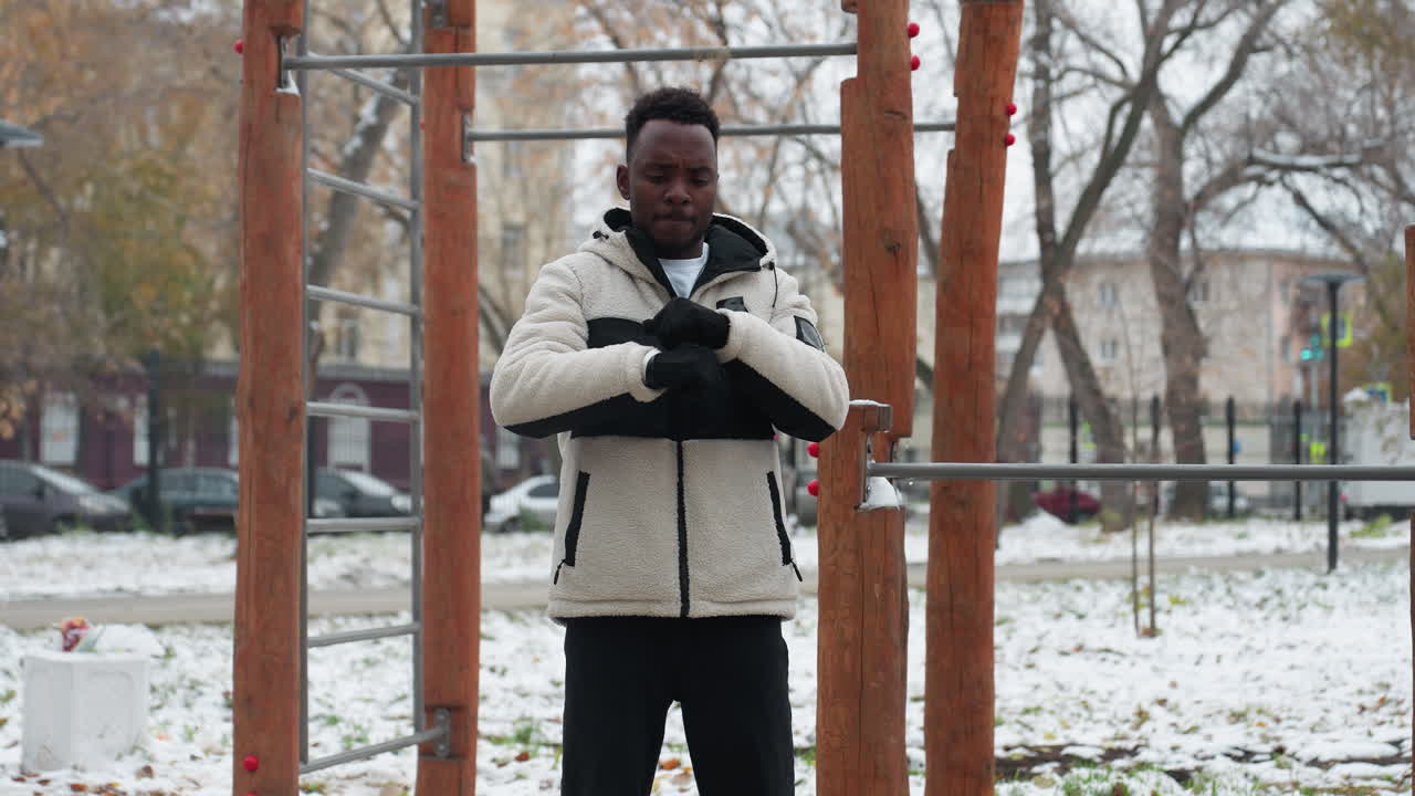 Uomo di colore che fa stretching all'aperto in inverno. Neve al suolo, attrezzatura da allenamento nel parco, attenzione all'attività fisica all'aperto nella stagione fredda con sfondo urbano