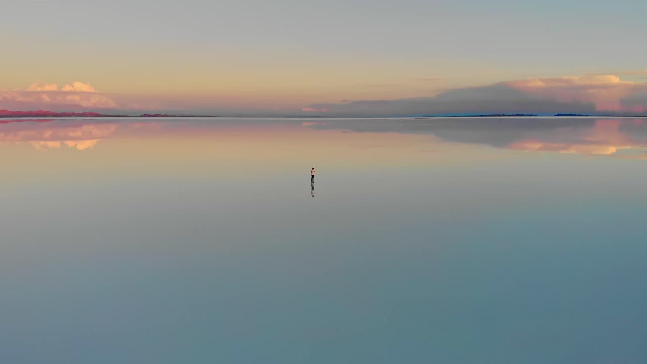 Aerial of a lone figure standing mirrored reflection of the world's largest salt flat at dusk in Uyuni Salt Flats , Bolivia