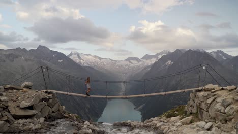 a blonde girl in a white dress walks from left to right across a suspension bridge