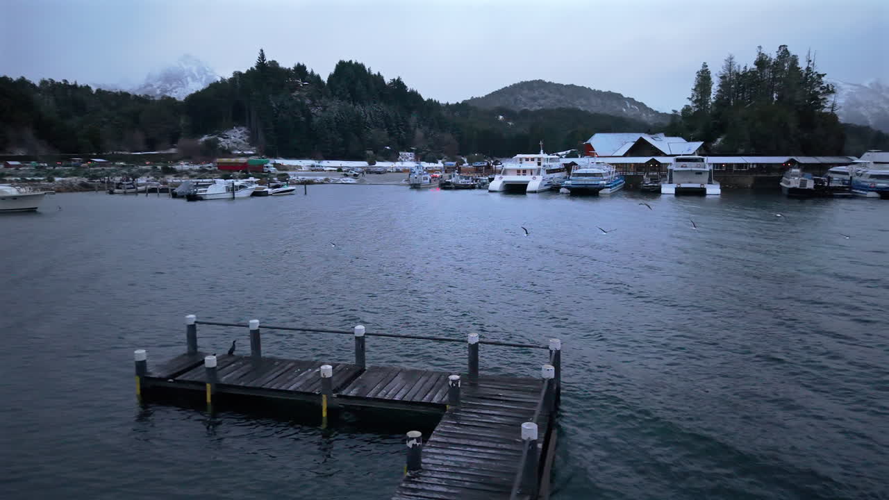 Drone descends toward Puerto Pañuelo marina on Lake Nahuel Huapi, Bariloche, Argentina. Snow covers roofs and hills, boats docked in cold waters, and mountains rise in the misty winter background.