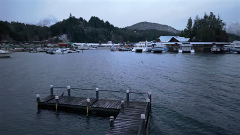 drone descends toward puerto pañuelo marina on lake nahuel huapi, bariloche, argentina. snow covers roofs and hills, boats docked in cold waters, and mountains rise in the misty winter background.