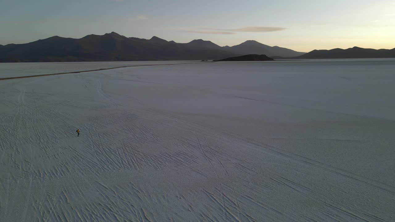 Tilted aerial of person walking on empty Uyuni salt flat with clear reflection and early evening light