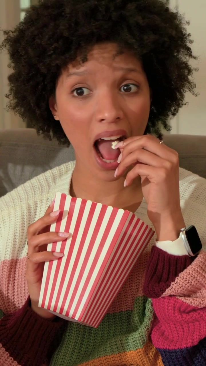 Video verticale: Donna che guarda, prende popcorn da un contenitore a strisce e reagisce alla TV sul divano.