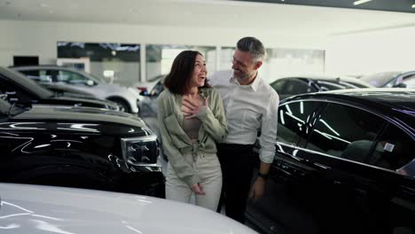 A-happy-middle-aged-man-with-gray-hair-in-a-white-shirt-opens-the-eyes-of-his-happy-brunette-girlfriend-with-his-hands.-I-present-her-with-a-white-car-that-he-bought-for-her-as-a-gift-at-a-car-dealership