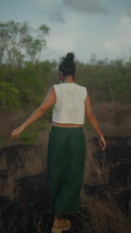 indian woman in green pants and a crop top walks across a charred, dry forest area. india